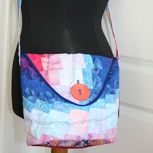 Könnte beinhalten: Eine Patchwork-Umhängetasche aus Stoff in den Farben Blau, Rosa und Weiß. Die Tasche hat einen großen, runden, orangen Knopf mit einem Schlüsselloch.