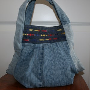 Könnte beinhalten: Eine blaue Jeans-Tragetasche mit einem dekorativen, bestickten Band mit roten, goldenen und blauen Pfeilen. Die Tasche hat einen breiten, flachen Boden und zwei Henkel.