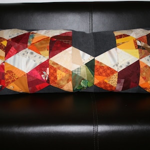 Könnte beinhalten: Ein langes, rechteckiges Kissen mit Patchwork-Muster. Das Kissen besteht aus verschiedenen Stoffen in Rot-, Orange-, Gelb- und Brauntönen. Das Kissen liegt auf einem schwarzen Ledersofa.