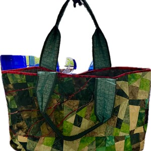 Op de afbeelding: Een groene en bruine patchwork-tas met rode stiksels en twee groene stoffen handvatten.