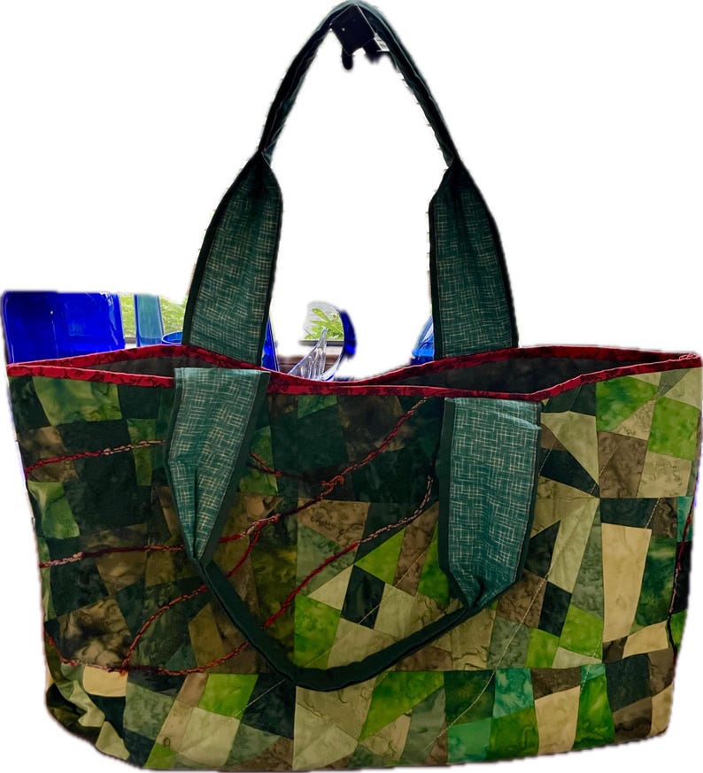 Op de afbeelding: Een groene en bruine patchwork-tas met rode stiksels en twee groene stoffen handvatten.