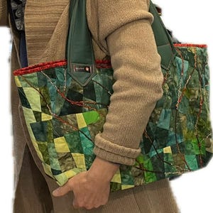 Op de afbeelding: Een groene, gele en blauwe patchwork tote bag met een groene leren band en een rode rand. De tas wordt vastgehouden door een persoon die een bruine trui draagt.