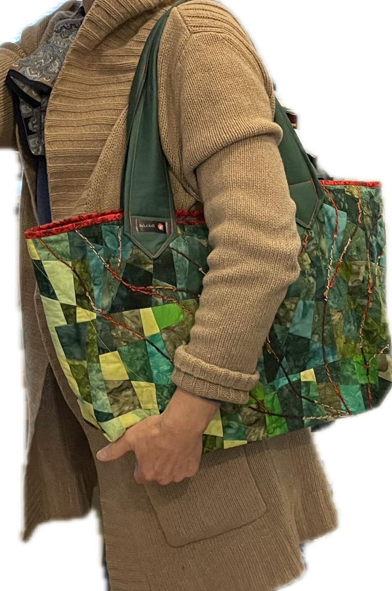 Op de afbeelding: Een groene, gele en blauwe patchwork tote bag met een groene leren band en een rode rand. De tas wordt vastgehouden door een persoon die een bruine trui draagt.