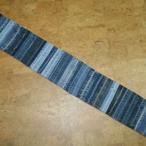 Puede incluir: Una bufanda larga y estrecha hecha de tiras de mezclilla azul cosidas juntas. La bufanda tiene un diseño de patchwork con diferentes tonos de azul.