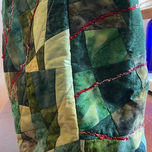 Op de afbeelding: Een groene en cr&egrave;mekleurige patchwork-tas met rode en roze stiksels. De tas is gemaakt van vierkante stukken stof die in een geometrisch patroon aan elkaar zijn genaaid.