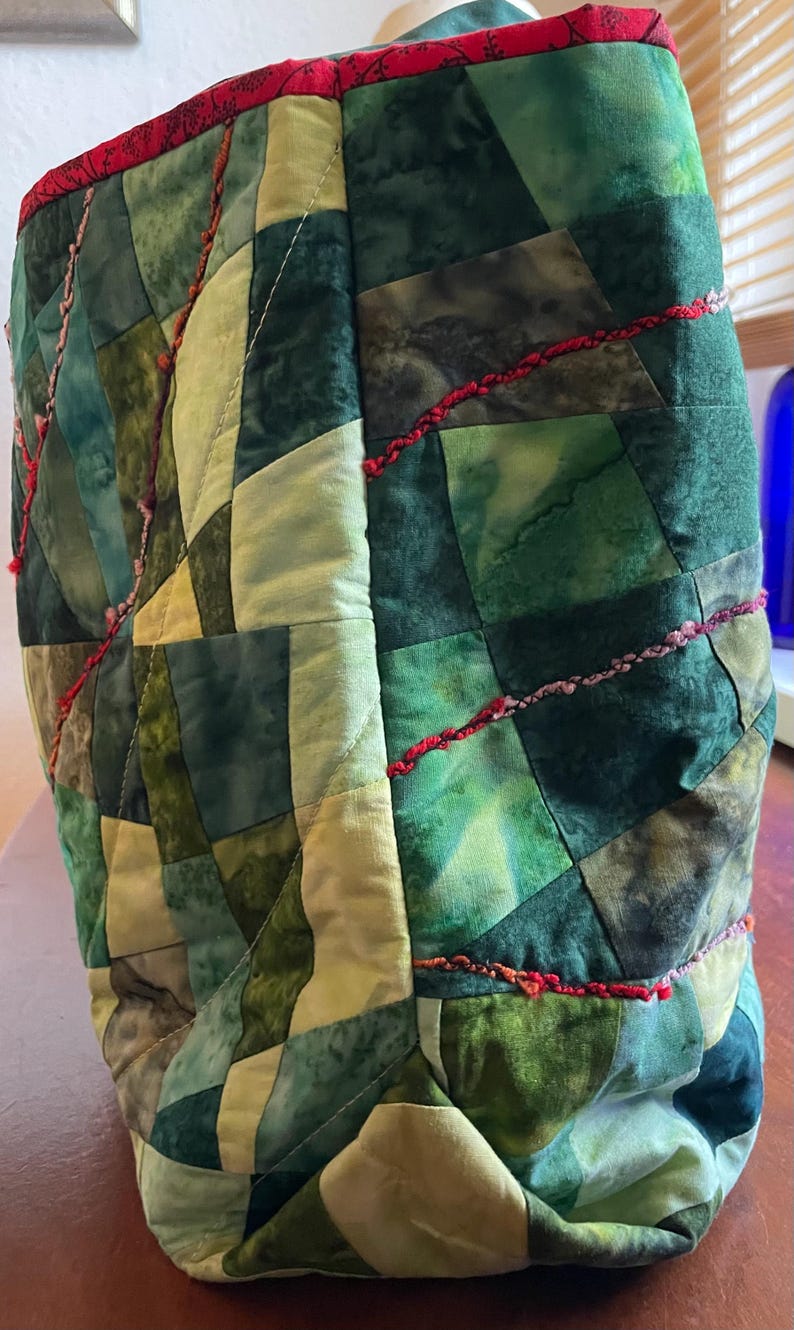 Op de afbeelding: Een groene en cr&egrave;mekleurige patchwork-tas met rode en roze stiksels. De tas is gemaakt van vierkante stukken stof die in een geometrisch patroon aan elkaar zijn genaaid.