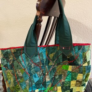 Op de afbeelding: Een grote groene en rode patchwork-tas met een gestikt ontwerp. De tas heeft een rode rand en twee groene hengsels. De tas staat op een houten stoel.