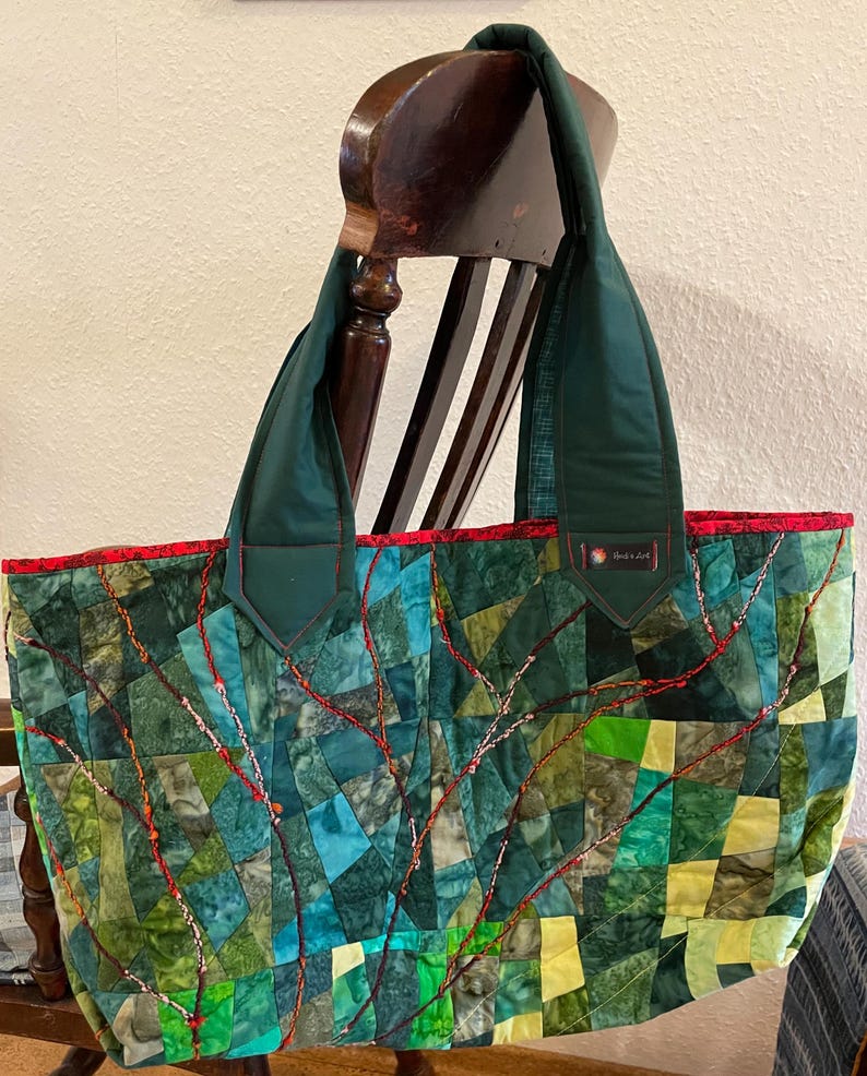 Op de afbeelding: Een grote groene en rode patchwork-tas met een gestikt ontwerp. De tas heeft een rode rand en twee groene hengsels. De tas staat op een houten stoel.