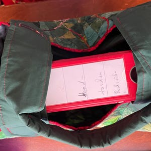 Op de afbeelding: Een groene stoffen tas met rode bies en een rode map erin. De map heeft een wit label met de tekst "Hand-fosch en Rucksooke".