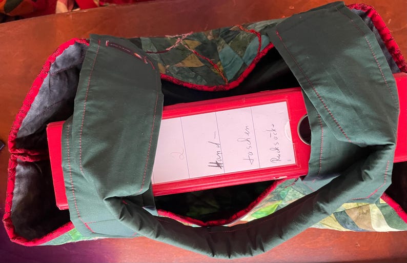 Op de afbeelding: Een groene stoffen tas met rode bies en een rode map erin. De map heeft een wit label met de tekst "Hand-fosch en Rucksooke".