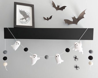 May include: A white felt ghost garland with black and white felt balls hanging from a black shelf. There are also black paper bats on the wall behind the garland.