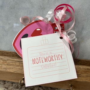 Può includere: Un blocco note rosa a forma di cuore con un biglietto bianco attaccato. Il biglietto ha linee rosa e rosse e il testo "You're a NOTEWORTHY Valentine!"