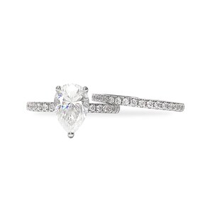 May include: A silver-toned ring set featuring a pear-shaped centre stone and a matching band, both adorned with small pavé-set stones along the shank. The pear-cut stone is held by prongs, and the bands are thin, metallic, and reflective in appearance.