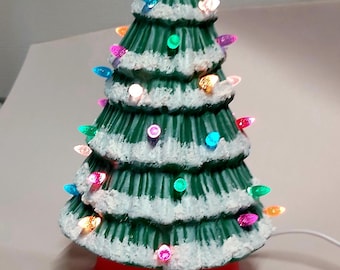 May include: A ceramic Christmas tree with a green body, white snow accents, and colorful lights. The tree sits on a red base and is topped with a clear star. The lights are multicolored and add a festive touch to the holiday decoration.