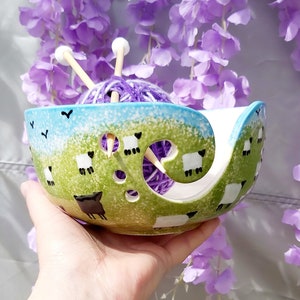 May include: A green and blue ceramic yarn bowl with a white sheep design. The bowl has holes for yarn to feed through and is holding a purple yarn ball and two knitting needles.