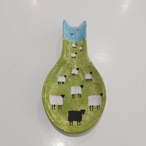 May include: A ceramic spoon rest featuring a hand-painted green pasture scene with multiple white sheep and one black sheep. The top section depicts a light blue sky with two small black birds. The piece has a curved handle and a wide, rounded base.