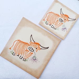 May include: Two ceramic tiles with hand-painted orange and brown Highland cow designs. Each tile features a cow with a flower and the text "UUUU" in a stylized font.