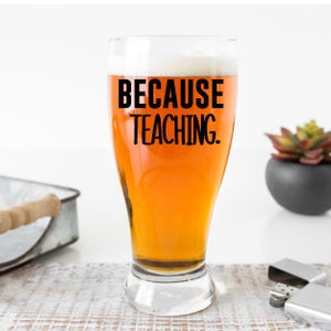 May include: A clear glass beer mug with the text "Because Teaching." printed in black on the front. The mug is filled with a light amber colored beer.