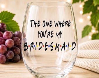 以下が含まれることがあります： The one where you're my BRIDESMAIDという黒い文字が書かれた透明なステムレスワイングラス。bridesmaidという単語の文字の間には小さなカラフルなドットがあり、紫色のブドウの房の前に置かれています。