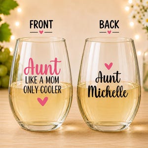 May include: Two clear stemless wine glasses filled with a light-colored liquid. The left glass features pink and black text reading Aunt LIKE A MOM ONLY COOLER with pink hearts. The right glass features black text reading Aunt Michelle with a pink heart.