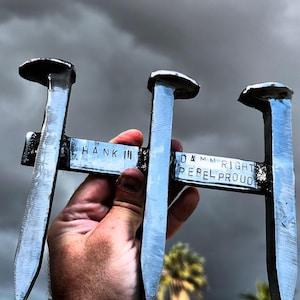 May include: A metal sculpture made from railroad spikes, held in a hand. The sculpture has the words "HANK III" and "DAMN RIGHT REBEL PROUD" stamped on it. The background is a cloudy sky.