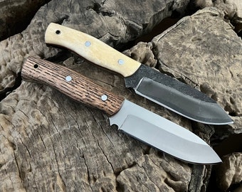 May include: Two knives with different handle materials and blade finishes. One knife has a brown wood handle and a silver blade, while the other has a white bone handle and a black blade. Both knives are on a wooden surface.