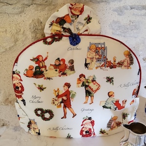 Può includere: Un copri-teiera bianco con un bordo rosso e un design natalizio festoso con bambini, Babbo Natale e ghirlande. Il design include il testo "Merry Christmas", "A Jolly Christmas" e "Greetings".