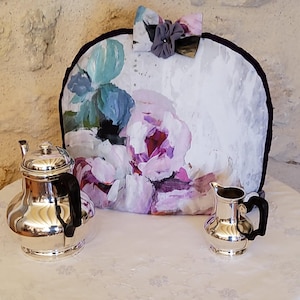 Può includere: Un servizio da tè in argento con una teiera floreale. Il set include una teiera, una lattiera e una teiera con un grande motivo floreale nei toni del viola, del blu e del bianco. La teiera ha un bordo nero e un fiocco in alto.