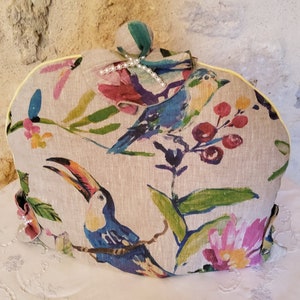 Puede incluir: Una funda para tetera beige con un patrón floral que presenta tucanes, pájaros y flores en tonos azules, verdes, rosas y amarillos. La funda tiene un ribete amarillo y una decoración de perlas en la parte superior.
