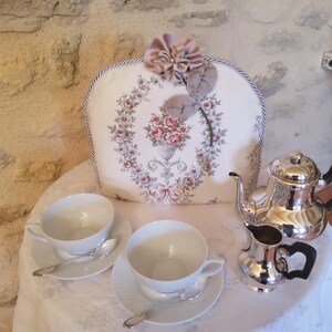 Peut inclure: Un service à thé sur une nappe blanche. L'ensemble comprend deux tasses et soucoupes blanches avec des cuillères, une théière en argent et un pot à crème assorti. Un chauffe-théières floral avec une fleur est posé derrière le service à thé.