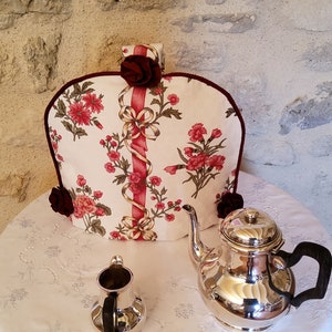 Puede incluir: Un cubre-tetera blanco con un patrón floral y una cinta roja. El cubre-tetera está sobre un mantel blanco con una tetera y una jarra de crema de plata.