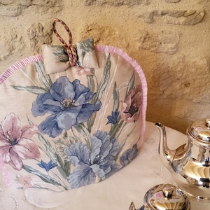 Può includere: Un copriteiera floreale con un bordo rosa e un fiocco. Il copriteiera è realizzato in tessuto beige con un motivo floreale in tonalità di blu, rosa e verde. Il copriteiera è appoggiato su una superficie bianca.