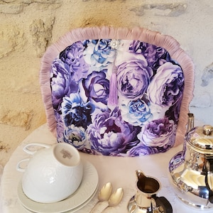 Può includere: Un copri-teiera floreale viola e blu con un bordo di frangia rosa. Il copri-teiera è appoggiato su una tovaglia bianca con una teiera d'argento, un creamer e una tazza da tè.