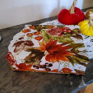 Peut inclure: Un plateau décoratif avec un motif sur le thème de l'automne, avec des champignons, des grenades et des feuilles d'automne. Le plateau est accompagné de trois citrouilles en velours rouge, jaune et vert. Le plateau est blanc avec un bord festonné.