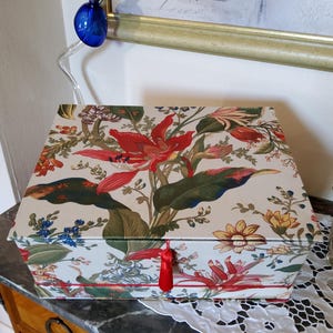 Könnte beinhalten: Eine rechteckige Box mit einem Blumenmuster aus roten, grünen und gelben Blumen auf cremefarbenem Hintergrund. Die Box hat eine rote Quaste und steht auf einer marmorgefliesten Oberfläche.