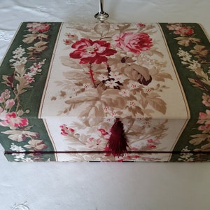 Könnte beinhalten: Eine Schachtel im Vintage-Stil mit einem Blumenmuster in den Farben Rosa, Rot, Weiß und Grün. Die Schachtel hat einen burgunderfarbenen Quasten.
