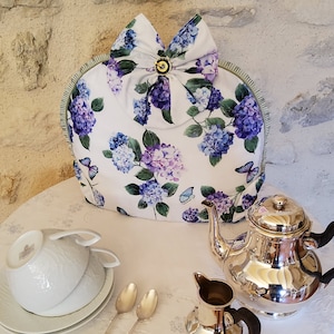 Può includere: Un copriteiera bianco con un motivo floreale di ortensie viola e blu e farfalle. Il copriteiera ha un bordo verde e un fiocco bianco con un bottone dorato al centro.