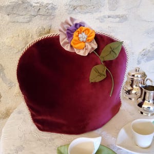 Può includere: Una teiera bordeaux con un'applicazione decorativa di fiori e foglie. Il fiore ha petali arancioni, viola e beige con un centro di strass. Sono visibili anche un servizio da tè in argento e una tazza da tè bianca.