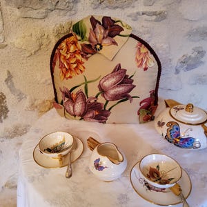 Può includere: Un servizio da tè con una teiera con motivo floreale, una teiera, una lattiera e due tazze con piattini. La teiera e la lattiera presentano disegni di farfalle. Il servizio da tè è su una tovaglia bianca.