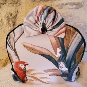 Può includere: Un copriteiera bianco con una stampa tropicale che presenta un pappagallo e foglie verdi. Il copriteiera ha un fiocco di tessuto abbinato in cima.