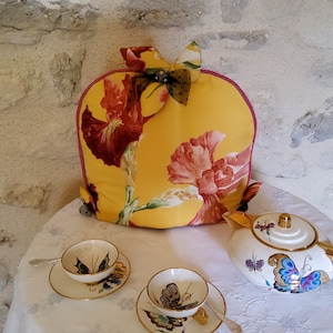 Può includere: Un copri-teiera giallo con un motivo floreale di fiori rossi e rosa e uno stelo verde. Il copri-teiera ha un fiocco fatto di un tessuto trasparente con un motivo verde e oro. Il copri-teiera è seduto su una tovaglia bianca con una teiera e due tazze da tè.