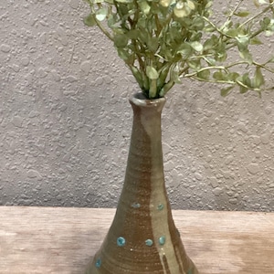 May include: A handmade ceramic vase with a brown and tan glaze, accented with turquoise dots and a turquoise base. The vase holds a bundle of faux greenery. The vase is displayed on a wooden surface.