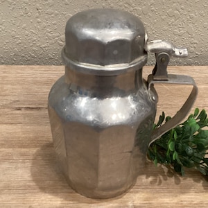May include: A silver metal creamer with a hinged lid and a handle. The creamer has a faceted design and is sitting on a wooden surface.