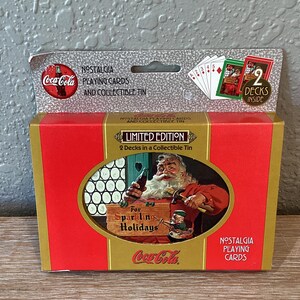 May include: A red and gold tin box with a Coca-Cola logo and the text "Limited Edition" and "2 Decks in a Collectible Tin". The tin features a Santa Claus character holding a bottle of Coca-Cola and a Christmas tree. The text "For Sparkling Holidays" is on a red and white sign.