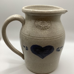 May include: A stoneware pitcher with a handle and spout. The beige-colored pitcher features a dark blue heart and paw prints design. The pitcher has the text "R.W. Miller" in a circular emblem.
