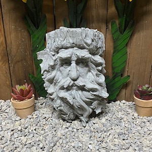 May include: A gray concrete garden sculpture of a bearded man's face with a leafy crown. Three yellow metal sunflowers with brown centers are mounted on a wooden wall behind the sculpture. The sculpture is sitting on a bed of gray gravel.