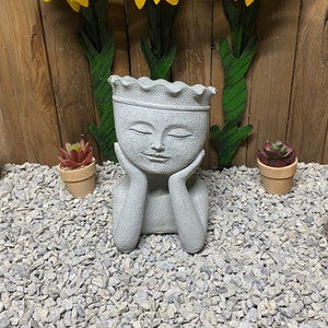 May include: A gray concrete planter shaped like a face with a crown on top. The planter is surrounded by small potted succulents and three yellow metal sunflowers.