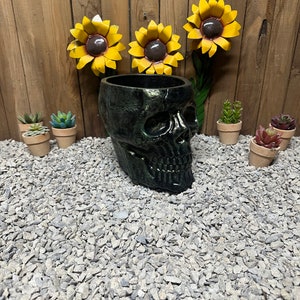 May include: A black skull-shaped planter with a green metallic finish. Three yellow metal sunflowers are attached to a wooden wall behind the planter. Small potted succulents are arranged in front of the planter on a bed of gray gravel.