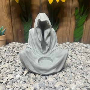 May include: A gray concrete statue of a hooded figure in a seated position. The statue has a hollowed-out area in the center, likely for a candle or small object. The figure is on a bed of gray gravel.