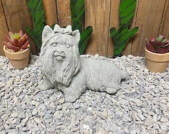 May include: A gray concrete statue of a Yorkshire Terrier dog wearing a pink bow. The statue is sitting on a bed of gray gravel.